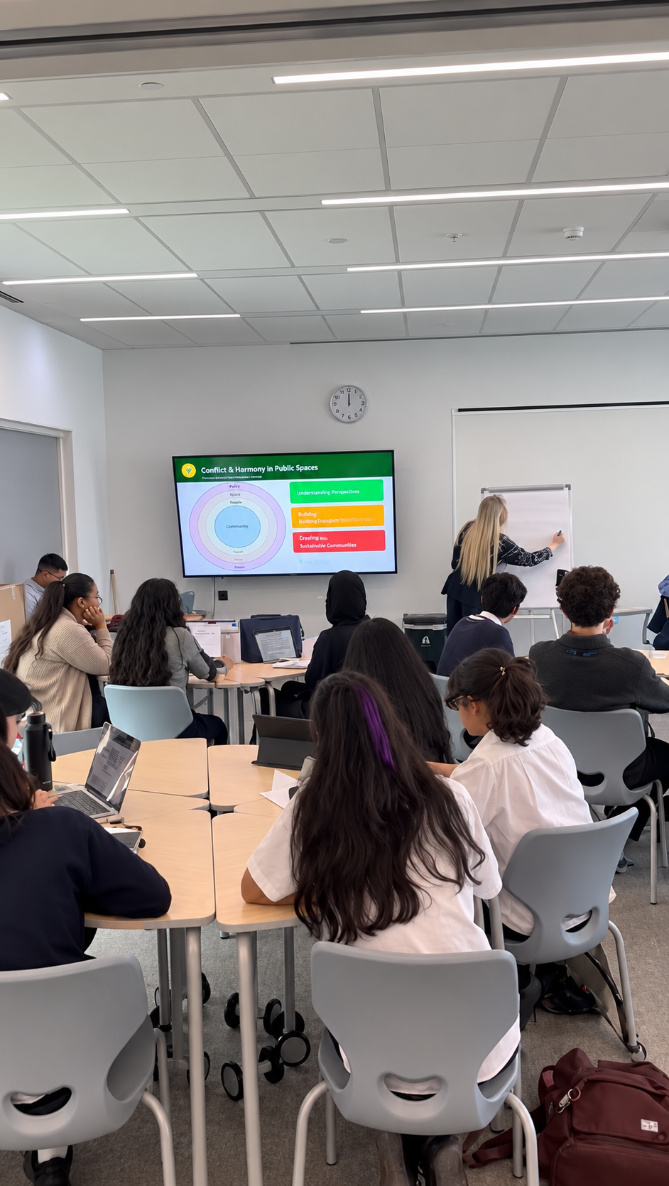 A teacher presenting to students with laptops in a modern classroom with a large wall screen